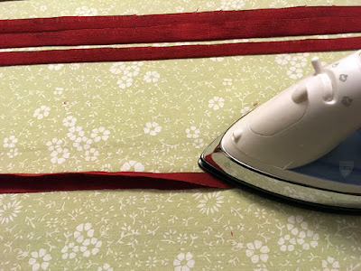 The nose of an iron pressing a narrow strip of red linen in half lengthwise on a pale green floral ironing board, with three other pressed strips arraged parallel at the far edge of the board.