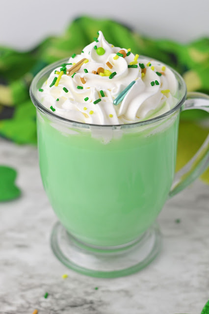 Green Hot Chocolate in a clear mug with whipped cream and sprinkles on top.