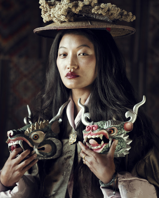 Uma butanesa branca, aparentemente jovem adulta. Tem cabelos pretos compridos. Na sua cabeça, uma espécie de chapéu enfeitado com flores brancas e pequenas. Pouco abaixo dos ombros, ela segura duas esculturas de cabeças de dragão.