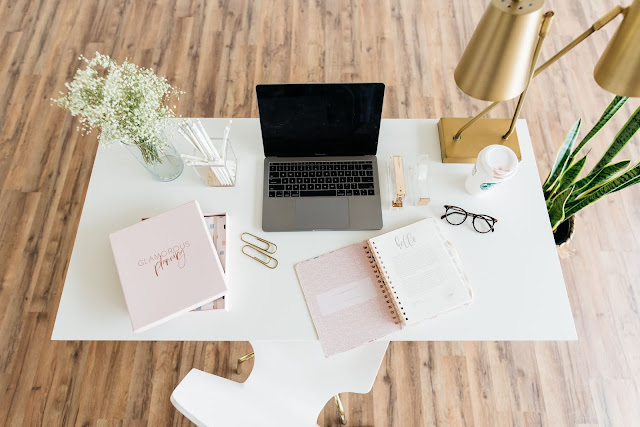 A laptop in the centre of a white desk. There is a vase of flaowers on the desk, along with notebooks and coffee cups.