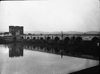 Slightly grainy black and white photograph of a stone bridge over a river. The still water dominates the image and reflects the arches and piers of the bridge which cuts across the centre of the image. There is a square stone tower centre left near the termination of the bridge and misty hills in the distance beyond the bridge.