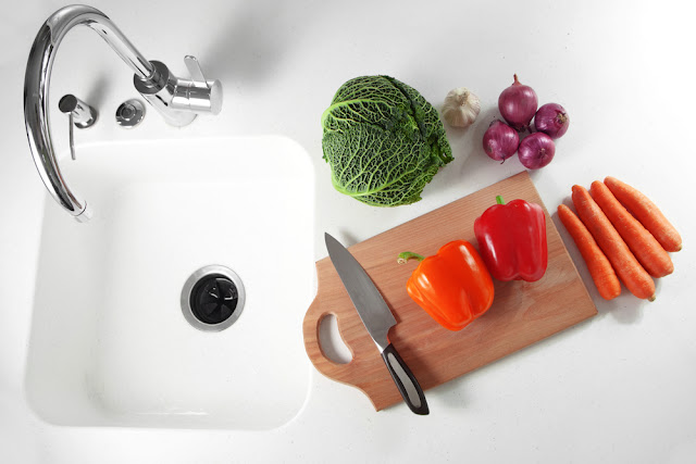 Stock photo showing a clean white kitchen sink and garbage disposal, with a cutting board featuring various colorful vegetables and a knife