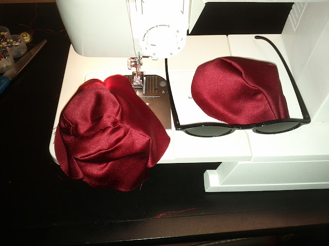 Red satin fabric starting to be sewn into a ball shape sits on a sewing machine with black sunglasses nearby.