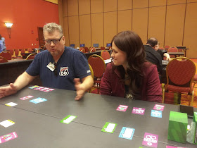 A man and a woman sitting at a table with a variety of the cards from the game arrayed on the table. The woman is listening intently to the man, who is speaking in a very animated manner with his hands out in front of him as if gesturing as he talks.