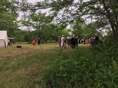 A view across a weedy fiedl at the edge of some trees, of a variety of people in medieval garb standing in groups chatting.