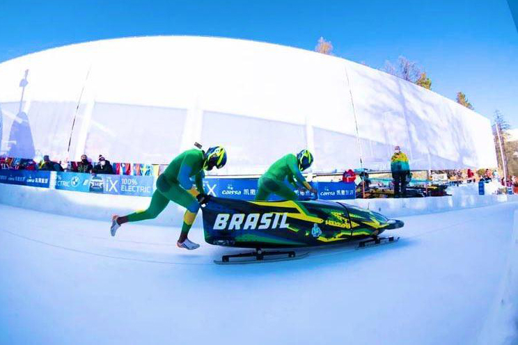 Dois atletas do Bobsled do Brasil, com roupas em detalhes verde e azul, empurram um trenó preto com detalhes azul e escrito Brasil em uma pista de gelo