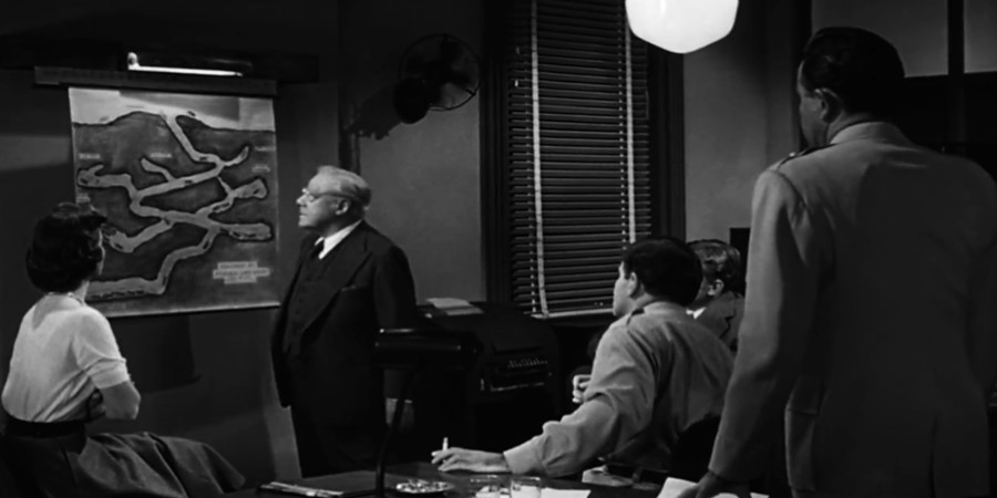 Fotograma en blanco y negro de la película "Them!" (1954). El Dr. Harold Medford, un hombre de cabello blanco y gafas, de pie frente a un dibujo de un hormiguero colgado en la pared mientras explica la expansión de las hormigas mutantes. Lo rodean cuatro personas (varios de los cuales llevan uniforme militar) que lo observan con atención, incluyendo una mujer sentada a la izquierda. La escena tiene un ambiente de oficina científica, con luz tenue, persianas y mobiliario austero, evocando la tensión y el estilo clásico de la ciencia ficción de los años cincuenta.