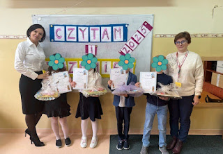 Na tablicy szkolnej napis : czytam i rozumiem, na jej tle stoi pani nauczycielka i pani bibliotekarka- organizatorki konkursu oraz dzieci - laureaci konkursu