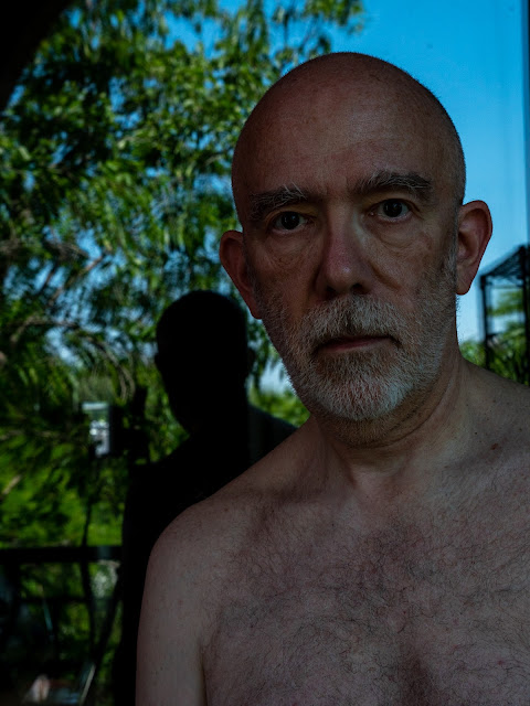 photograph of an older white male with a blue sky and green leaves reflected in a window behind him. His shadow on the glass is visible.