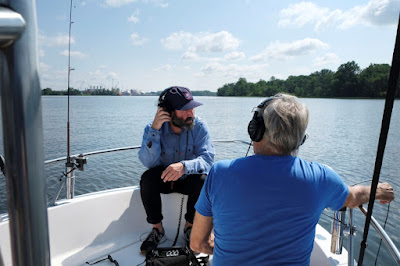 Two men in blue shirts sit in the front of a fishing boat on the Delaware River wearing headphones. One is holding a microphone cable over the side of the boat.