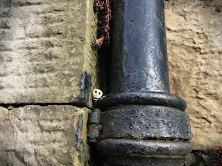 A close up photo showing a black drainpipe against a grey stone wall.  A small ceramic skull (Skulferatu 94) sits in a gap between the wall and the drainpipe.  Photo by Kevin Nosferatu for the Skulferatu Project.