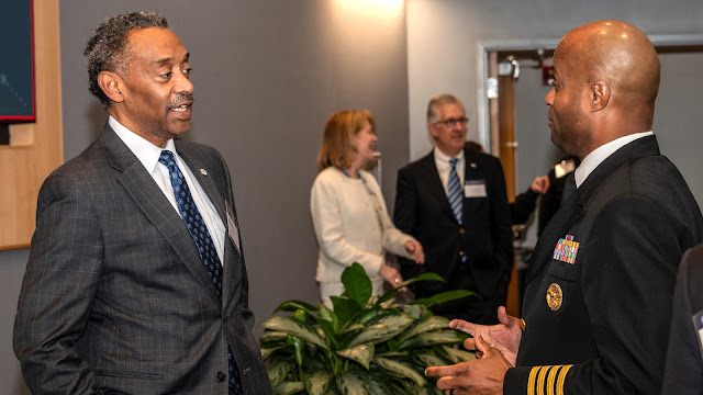 A group of people conversing in a professional setting. To the left, a man in a plaid suit is smiling and talking, while on the right, a man in a navy military uniform with medals is actively gesturing. Others in business attire are in the background, engaging in discussions.