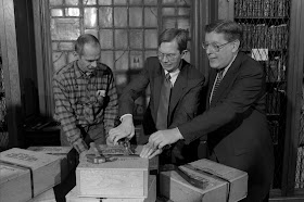 A photograph of three men opening a wooden box.