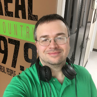 The picture:  a selfie of me, wearing a green shirt, standing before the logo of the radio station I used to work at