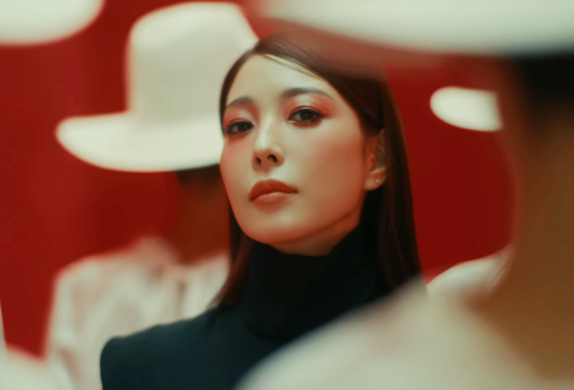 BoA shot in close-up, looking into the camera, wearing light red eye shadow. Stood amongst a group of women all wearing white white brimmed hats.