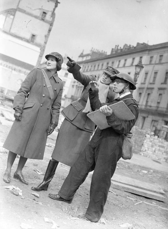 Women of the WW2 MTC - Miss Winifred Ashford points something out to an Air Raid Precautions (ARP) Warden with a clipboard, as Mrs Pat Macleod looks on. The Warden is based at Paddington ARP station. Behind them can be seen an empty space that once held houses before they were destroyed in an air raid.