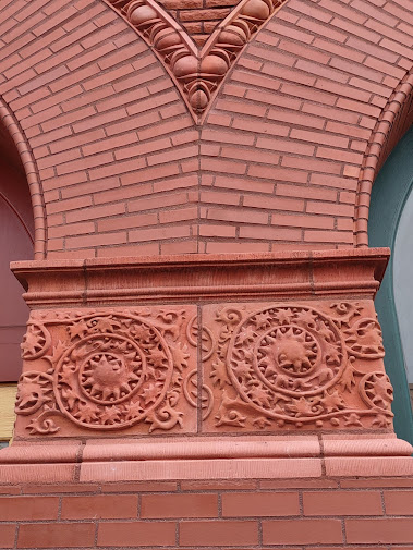 Photo: detail of a red brick building. Two arches meet in the center, on top of a horizontal section with two leafy vines (also in red plaster or brick) and a sort of plinth of horizontal red brick.