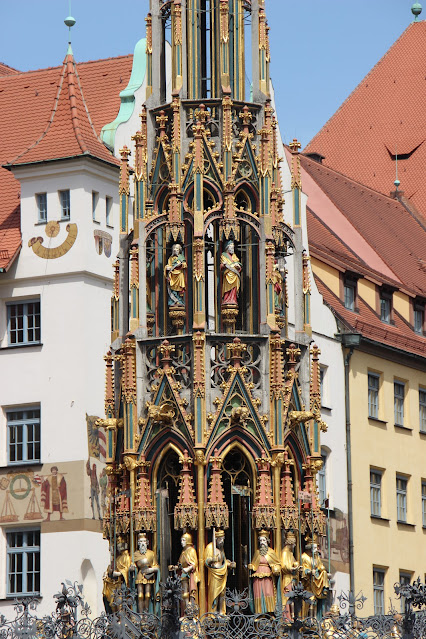 Colourful statues on a tall elaborately decorated pillar with  surrounded by an ornamented iron grille at its base in front of red-roofed white- and yellow facades of period properties.