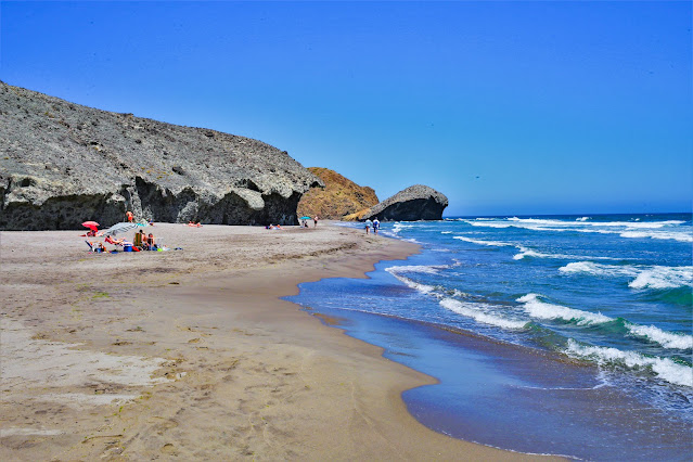 Orilla de playa de arena fina, con las olas del mar llegando y un paisaje rocoso de origen volcánico de fondo