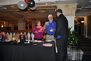 Picture of Annie and her Husband standing behind the ticket table, they are talking to a tall guy in a black suit, his back is to the camera