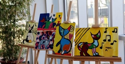 There are four colorful artistic paintings, each on its own paint stand, and all featuring a cat in them.