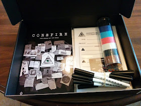 The Conspire Deluxe Set. A flat black box, propped open to reveal the Conspire rulebook, with artwork depicting a typical conspiracy theorist's bulletin board, showing the conspire logo, an eye in a triangle, in the centre. Also in the box is a tube containing poker chips in several different colours, a bundle of dry-erase markers, laminated role sheets, and a Cherry Picked Games business card.