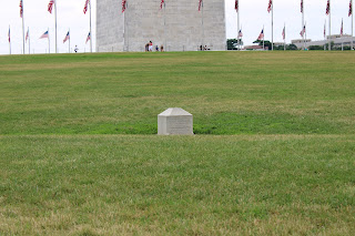 a small stone monument in a grassy area. the base of the washington monument can be seen in the background