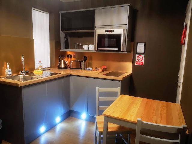 The interior of Bede Serviced Apartments, Coventry. The image shows an L shaped kitchen area with stainless steel sink, kettle and toaster. Oak effect worktops and grey gloss kitchen units. There are blue LED light strips along the bottom of the kitchen units and a bronze coloured splashback. There is a small hob set into the worktop and a microwave above it. To the right is a fire blanket for emergency use and in front of that there is a small dining table and chairs.