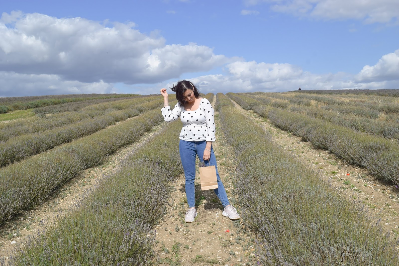 A photograph of me posing in the middle of the lavender field wearing a white and black polkadot long sleeve top and blue jeans. I am looking down with one hand in my hair and holding a brown paper bag with lavender in with my other hand.