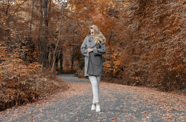stylish blonde woman standing in the middle of a trail with autumn clothes and fall background