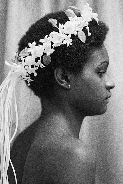 Fotografia, em preto e branco, contendo uma mulher negra, de costas, com cabelos curtos e usando uma grinalda de flores brancas.