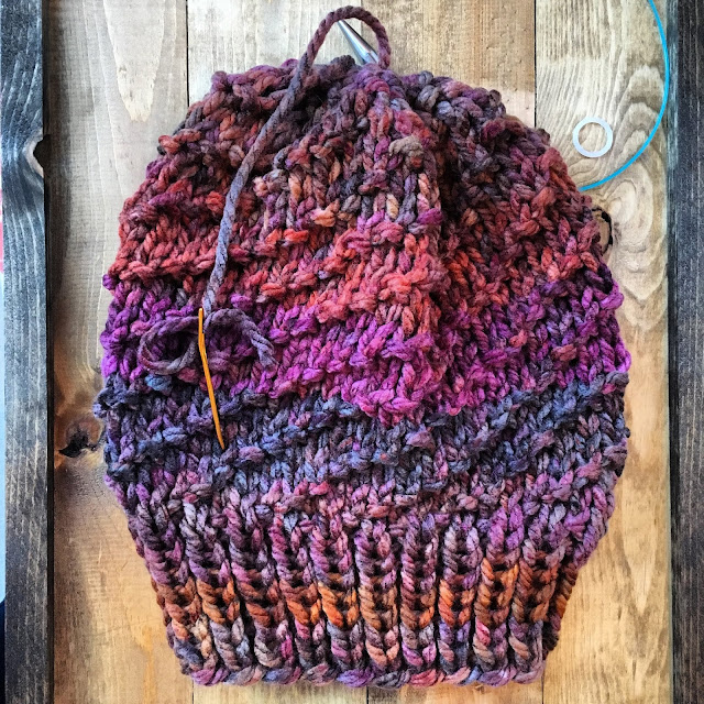 Flat shot of a warm earthy pinks and browns chunky knit hat with bumps lined up in diagonal lines. It lies on a wood surface. A gold yarn needle has the tail threaded through, waiting to be woven in.