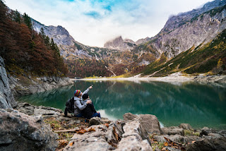 Two people sat beside a lake in forested mountainous terrain. One points out something to the other.