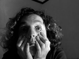 A playful black and white headshot of Poet and Performer Caroline Reid taken inside. She is using her fingers to pull down the skin on her cheeks, which distorts her face. One eye is in shadow, the other appears googly and oversized. Caroline is wearing a dark top. Her curly hair comes down to her shoulders.