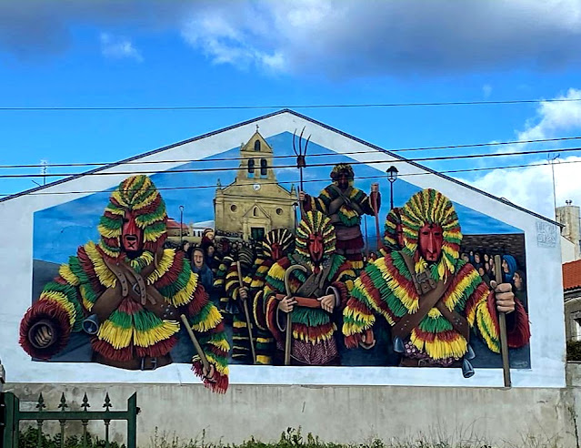 pintura numa parede branca de vários caretos de Podence com a igreja da aldeia ao fundo