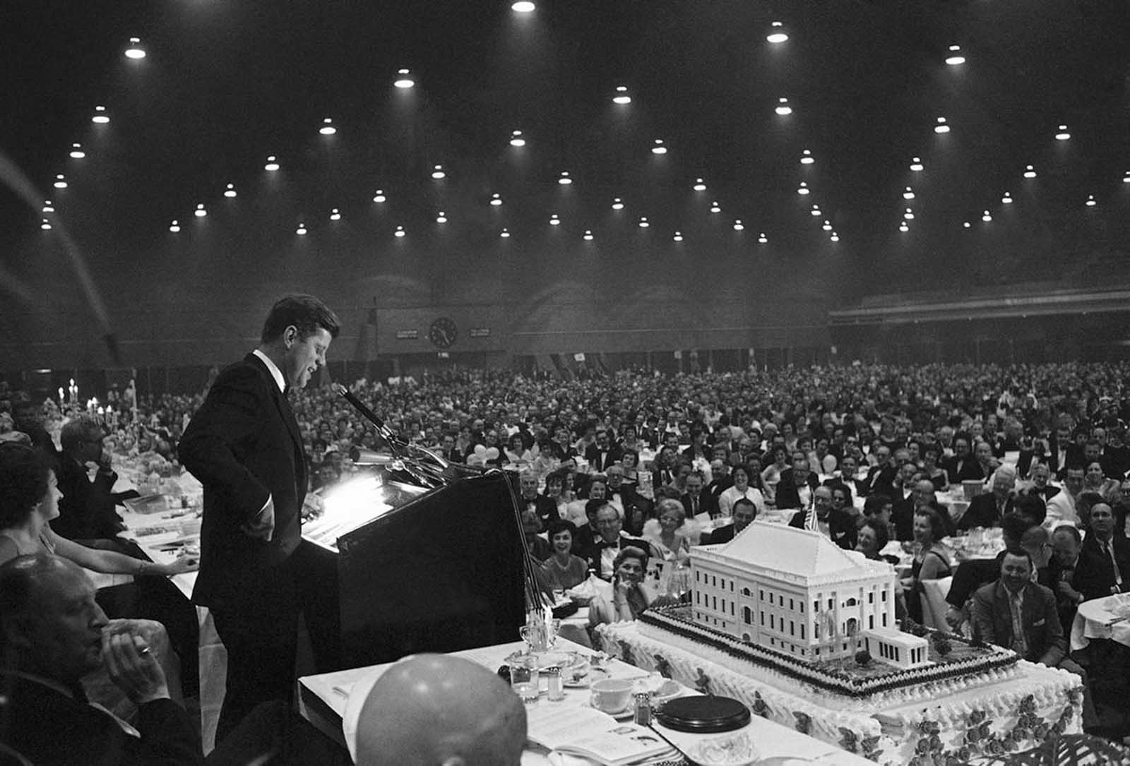 President Kennedy smiles as he delivers a joking remark to the 6,000 attending the $100-a-plate Democratic dinner on May 27, 1961 in Washington?s National Guard Armory, to help him celebrate his 44th birthday. The cake in foreground weights 1.5 tons and is topped by a replica of the White House.