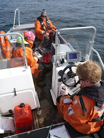 Kolme pelastautumispukuista henkilöä veneessä
