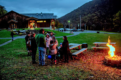 Group of adults and children next to picnic tables and a bonfire. The PA Lumber Museum visitor center, gray clouds, and Denton Hill are in the background.