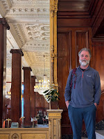 Man standing to the right of a giant, gilt mirror, which is reflecting a colonnade.