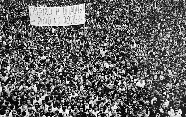 A fotografia é toda composta por uma multidão de manifestantes e há um cartaz e nele está escrito “abaixo a ditadura, povo no poder”.