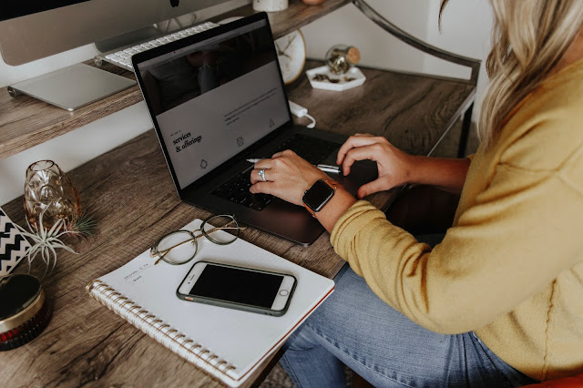 Woman working at a wooden desk on a laptop. Beside her is a notebook, a mobile phone and glasses