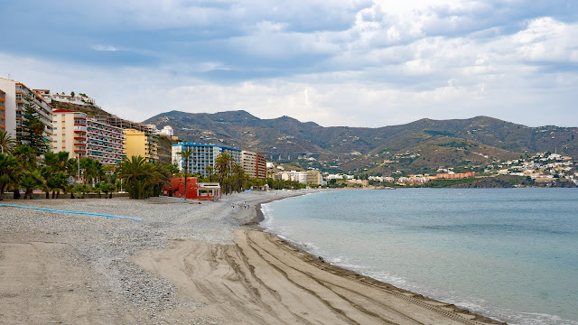 Playa con edificios a su espalda, montañas al fondo y las azules aguas del mar a su frente.