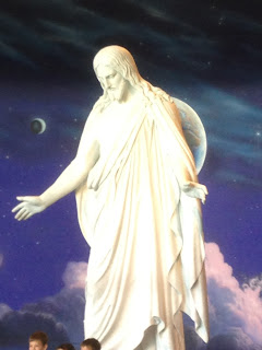 A white marble statue of Jesus Christ with open arms. A mural of stars and clouds is in the background. The tops of kids' heads peek into the bottom of the picture, they come up to his knees.