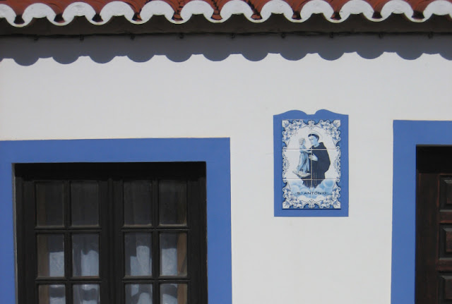 Fragment fasady jednego z domków w Odeceixe. Ściana pomalowana jest na biało. Drzwi i okna obwiedzone są niebieską linią. Niewielki obrazek z płytek ceramicznych malowanych w błękitach z przedstawieniem św. Antoniego Padewskiego dekoruje ścianę.