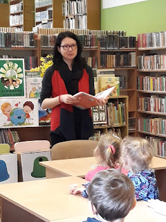 Pani bibliotekarka opowiada dzieciom o ochronie środowiska. Tło: sala biblioteczna