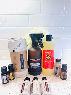 A neatly arranged display of dōTERRA's cleaning products on a light wooden surface. From left to right: On Guard Cleaner Concentrate in a clear bottle, Abode Microfiber Cloths stacked beside, a row of Abode Multi-Purpose Surface Cleaner Concentrate bottles, Abode Multi-Surface Spray Dispenser, and essential oil bottles of Purify, Lemon, and Wild Orange. The background is softly blurred, emphasizing the vibrant labels and clean design of the products.