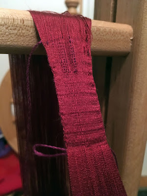 A close-up of a very small, rather rough band of deep garnet weaving, with the warp threads fanning over the wooden bar at the top of the photo and as they descend out of frame at the bottom. The top of the woven section is rippled and loose, but the lower portion is snug, with slight darker shadows at intervals and a loop of purple thread hanging left from the very bottom of the woven section.