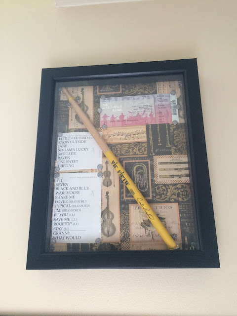 Photograph: A 3D picture frame with a background of printed instruments. Displayed are a set list, fan club ticket, and drumstick.