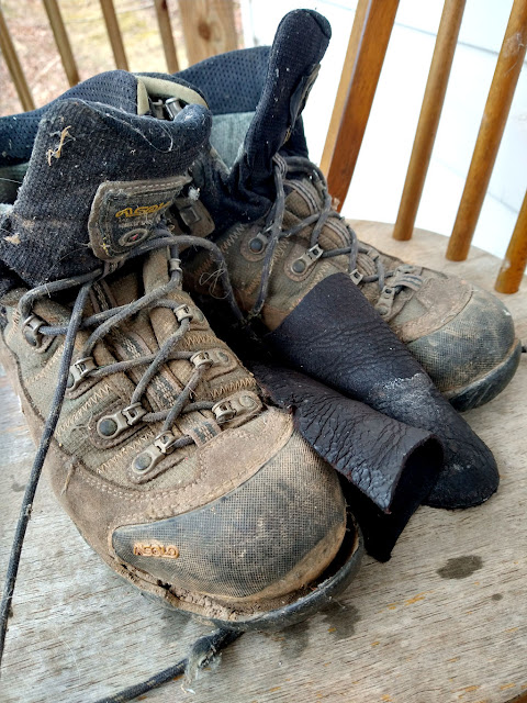 A pair of muddy hiking boots sitting on a weathered wooden chair waiting for disassembly with some previously salvaged shoe-leather sitting between them. The salvaged leather has darkened from all the oil used to condition it.