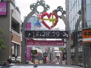 Gate at the start of a wide street of shops. The gate is shaped like an upside down U. There is a balloon scultpure placed on it of a woman with a bunny head holding a large red heart. Below the balloon scultpure is a large digital display of the time. Below the digital display is a pink banner with Japanese text on it. Inside the arch the shop signs are along the street.